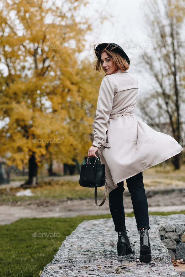 stylish woman in trench coat and hat walking by autumn park and turning back