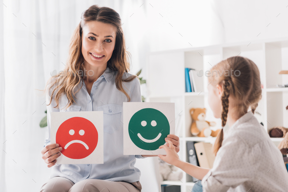 smiling psychologist showing happy and sad emotion faces cards to child