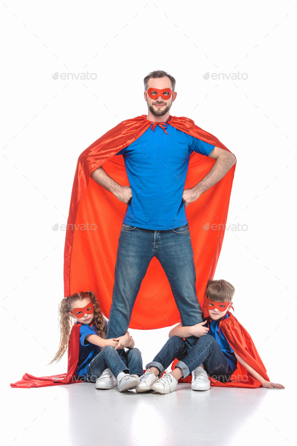 super dad standing with hands on waist and looking at camera while kids hugging his legs isolated on