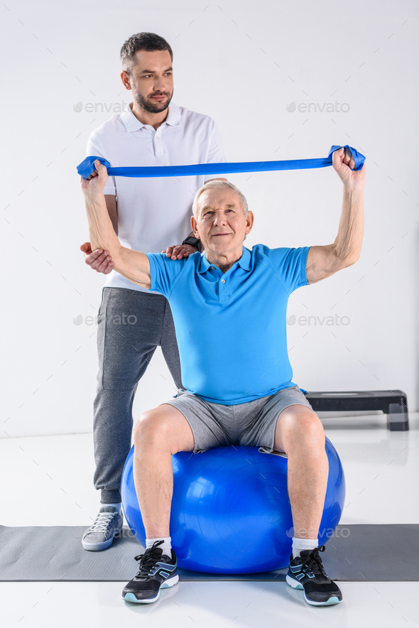 rehabilitation therapist assisting senior man exercising with rubber tape on fitness ball