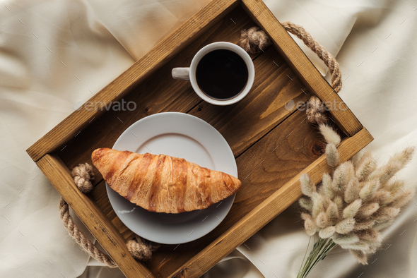 top view of coffee and croissant on tray with lagurus ovatus bouquet on beige cloth, breakfast in