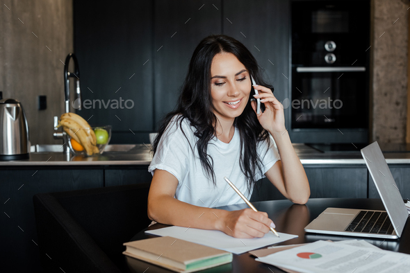 smiling woman talking on smartphone and working with laptop and business documents on kitchen during