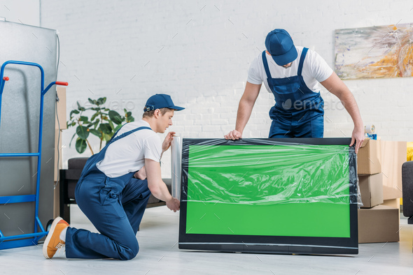 two movers in uniform using roll of stretch film while wrapping tv with green screen in apartment