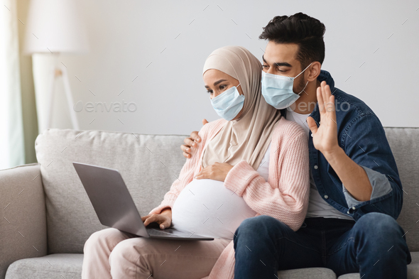Pregnant muslim couple wearing medical masks making video call with laptop at home