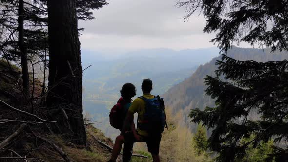 Aerial Flight Over Couple Sitting on Cliff Edge. Young couple hiking on top of the mountai. alt