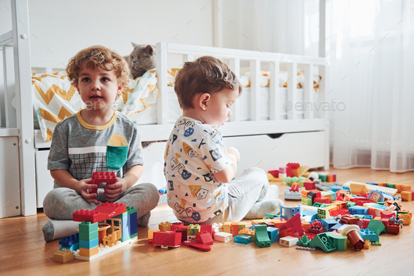 Two little boys have fun indoors in the bedroom with plastic construction set. Cat sits behind them