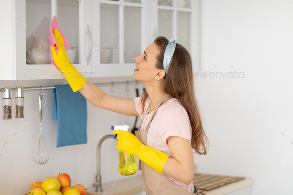 Lovely millennial housemaid wiping kitchen cabinet, performing house cleanup