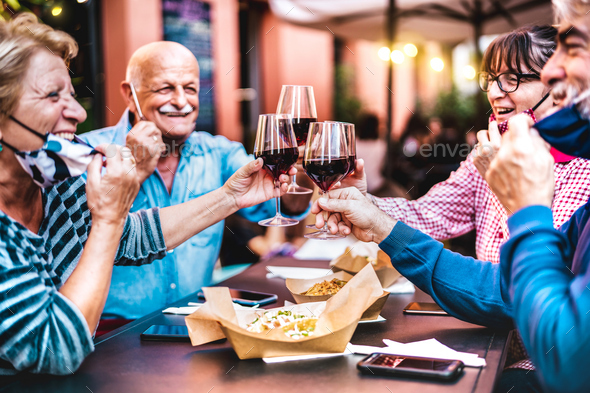 Senior people toasting wine at restaurant bar wearing open face masks