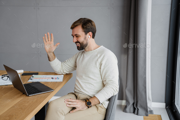 Handsome pleased man waving hand while working with laptop