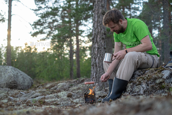 Man roasts a sausage over a fire in a metal camp stove and holds a mug of hot drink in hands