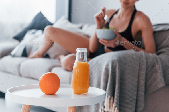Young woman with slim body shape in sportswear sits on sofa and eats healthy diet food
