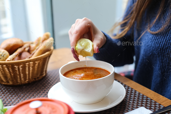 side view woman squeezes lemon into chicken soup in a cup with bread on the table