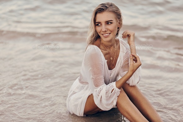 Gorgeous blond woman with perfect tan body lying on send near water in white dress.