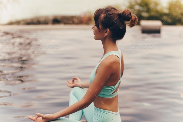 Caucasian woman meditating in lotus pose near pool during yoga retreat in tropical island.