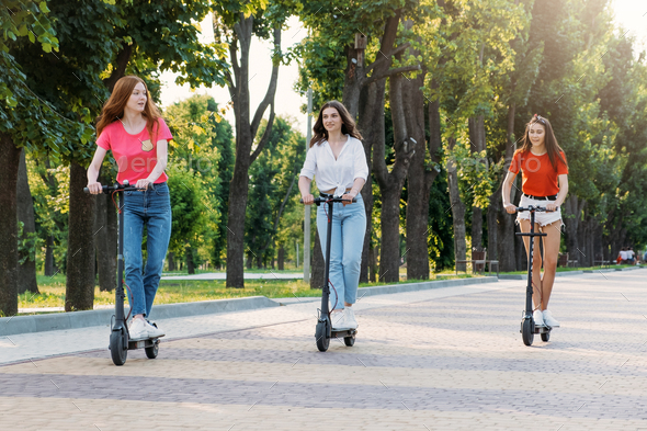 Three young girl friends on the electro scooters having fun in city street at summer sunny day