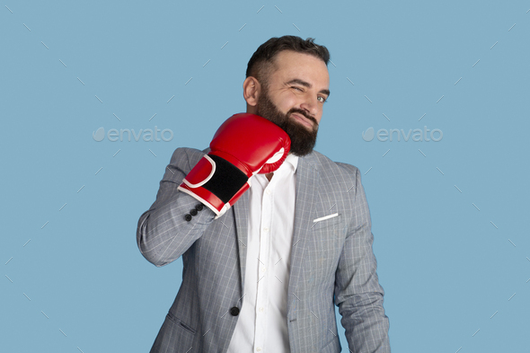 Young bearded businessman hitting himself in face with red boxing glove, blue studio background