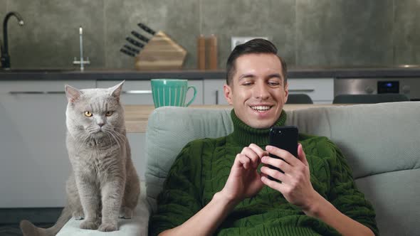 Young Man Uses a Smartphone and Lies on the Couch with a Domestic Cat in a Cozy Home alt