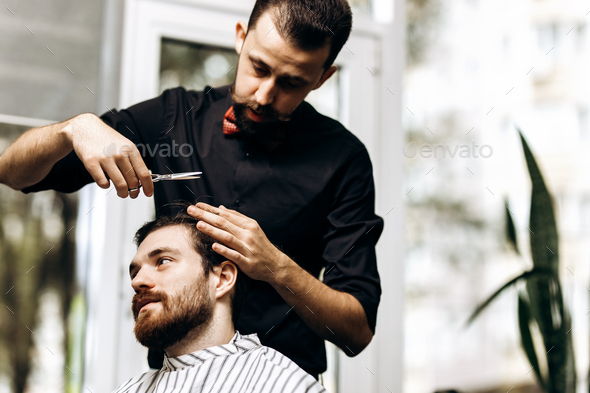 Barber dressed in a black shirt with a red bow tie makes a stylish hairstyle to young man in a