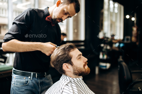 Barber with mustache dressed in a black shirt with a red bow tie makes a stylish hairstyle to young