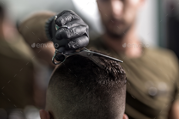 Barber in black gloves cuts with scissors hair on top of head of dark-haired man at a barbershop