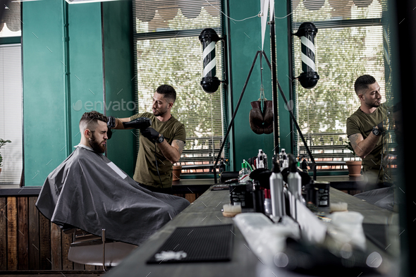 Young man with a beard sits in the chair in front the mirror at a barber shop. Barber dries mens