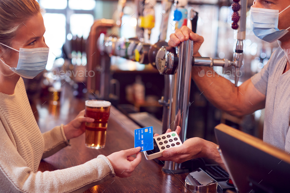 Female Customer Wearing Mask In Bar Making Contactless Payment For Drinks During Health Pandemic