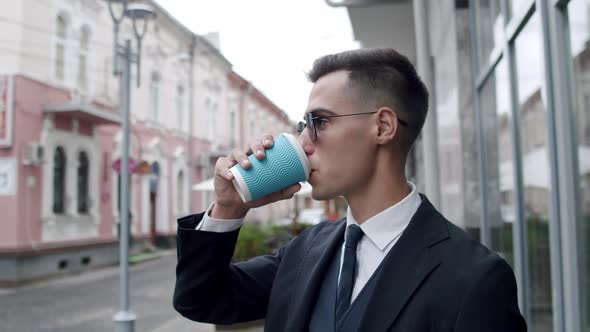 Elegant Man in Suit with Dark Sunglasses Drinks a Cup of Coffee and Looks Around alt