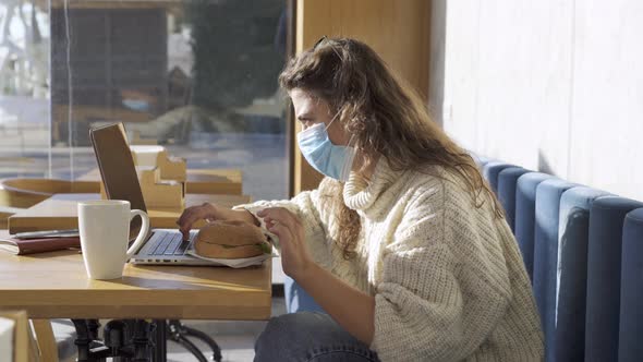 Woman with Mask Eats Bagel Burger Working on Laptop in Cafe