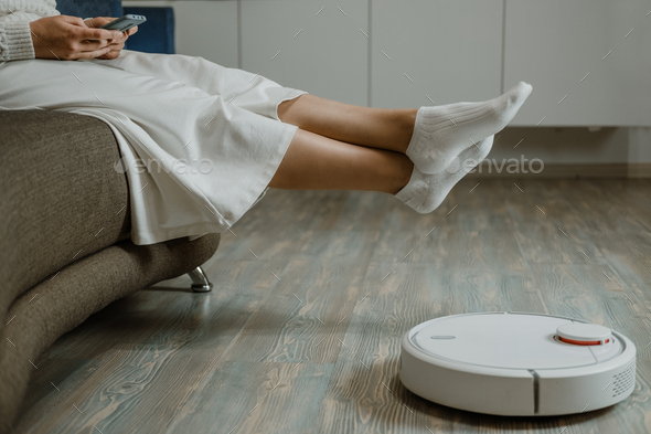 Robotic Vacuums, Robot Mops. Smart home. Robotic Vacuum Cleaner while Woman Relaxing on sofa