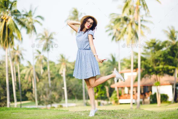 Young girl wearing sky-blue dress is standing on one leg on tip-toes in a park. Girl has straw hat