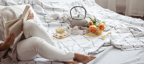 Woman reads in bed with tea, teapot and bouquet of flowers.