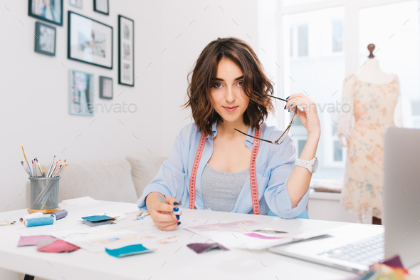 A brunette girl is sitting at the table in the workshop. She has blue shirt and creative mess on the