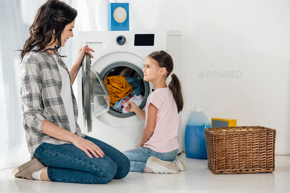 daughter in pink t-shirt and mother in grey shirt sitting on floor near washer with clothes in