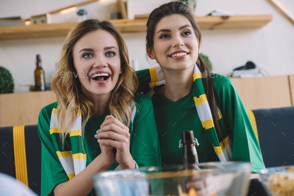happy female football fans in green t-shirts and scarf gesturing by hands during watch of soccer