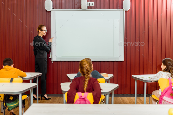 smiling teacher pointing at interactive whiteboard and looking at schoolkids