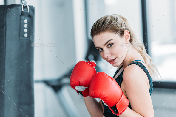 attractive young sportswoman in boxing gloves looking at camera in gym