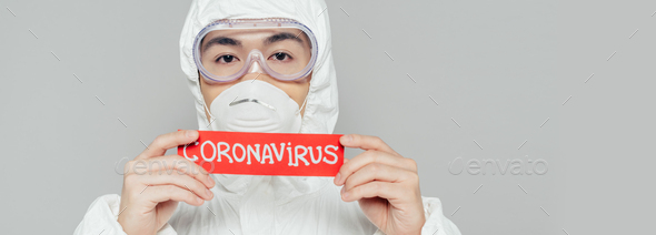 panoramic shot of asian epidemiologist in hazmat suit and respirator mask holding warning card with