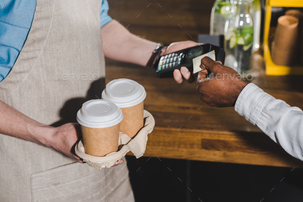 cropped shot of client paying by credit card and barista with terminal and coffee cups in hands