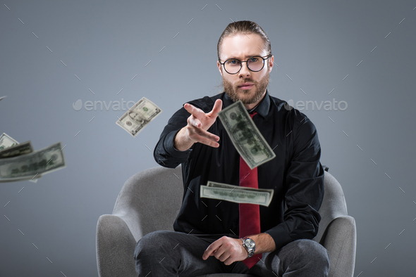 Young successful businessman in glasses sitting in armchair while throwing money to camera, isolated