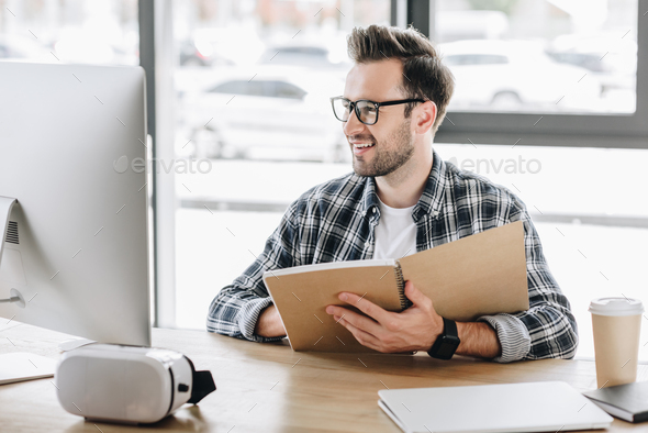 smiling young man in eyeglasses writing in notebook and using desktop computer at workplace