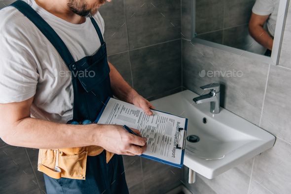 partial view of male plumber with toolbelt writing in clipboard near broken sink in bathroom