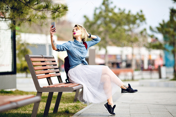 Young cute blond girl with short hair and bright pink lips sitting on a wooden bench making a selfie - Stock Photo - Images