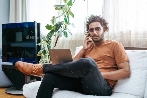 Handsome indian guy dressed in casual clothing sits on sofa with laptop