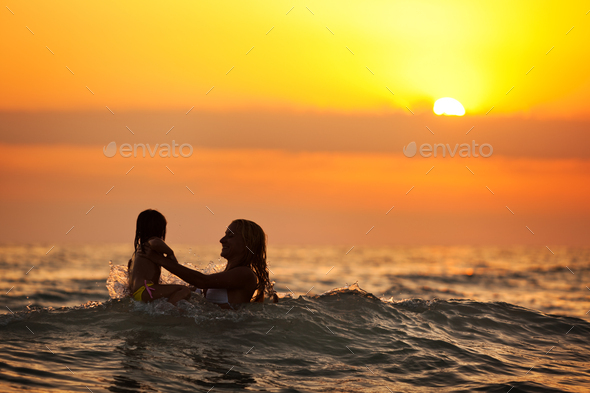 Young mother standing in water and playing with her daughter at sunset