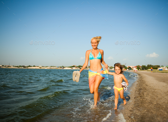 Young mother and her small daughter running on water edge and smiling
