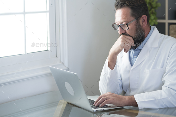 Mature doctor sitting in office. Adult doc in white coat write and work at desk with laptop