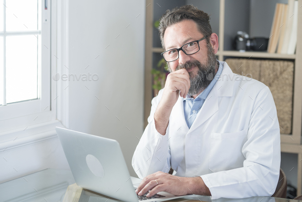 Smiling old male doctor in glasses and white uniform sit at desk in hospital work on laptop