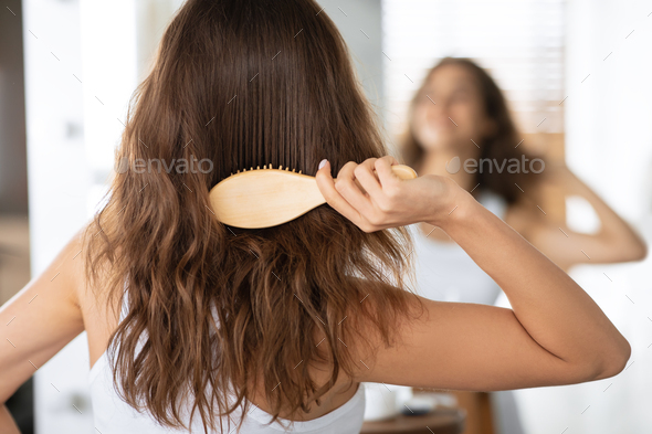 Woman Brushing Hair With Hairbrush Standing Near Mirror Indoor, Back-View