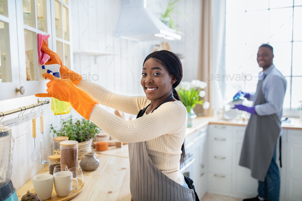 Happy black lady cleaning kitchen cabinet, her husband washing dishes on background