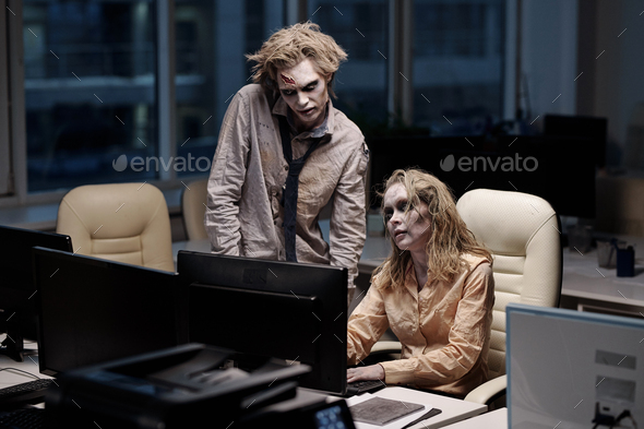 Young spooky zombie businessman standing next to female colleague sitting in front of computer